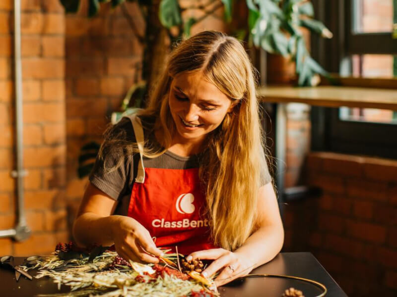 A smiling woman in an apron builds a Christmas wreath out of dried flowers